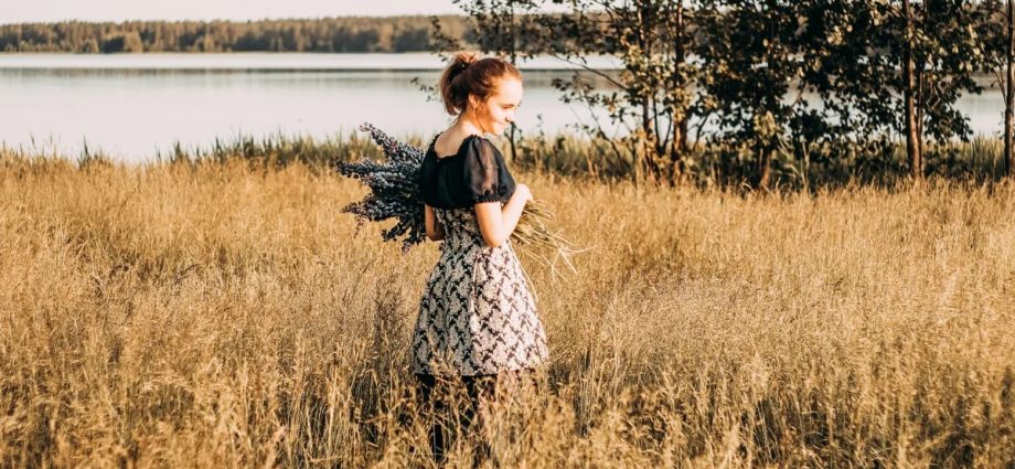 A young woman stands in a sunlit field by a lake, holding lavender flowers and smiling.