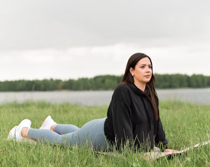 Woman lying on a yoga mat in a grassy field by a lake, promoting relaxation and wellness.