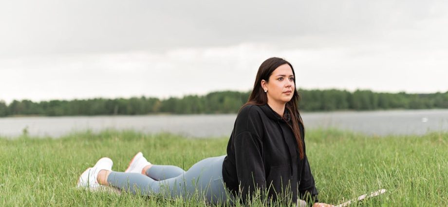Woman lying on a yoga mat in a grassy field by a lake, promoting relaxation and wellness.